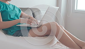 Young asian woman sitting on the bed checking social apps and working on laptop computer in the bedroom while working from home.