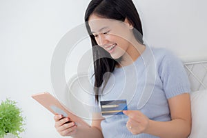 Young asian woman sitting on bed using digital tablet shopping online with credit card in the bedroom at home.