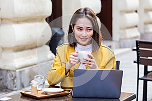 Young Asian woman sitting in coffee shop at wooden table, drinking coffee and using smartphone.On table is laptop. Girl browsing