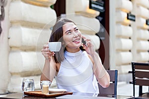 Young Asian woman sitting in coffee shop at wooden table, drinking coffee and using smartphone.On table is laptop. Girl browsing