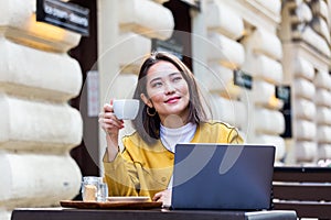 Young Asian woman sitting in coffee shop at wooden table, drinking coffee and using smartphone.On table is laptop. Girl browsing