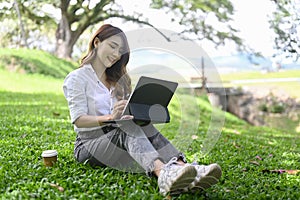 Young asian woman sitting on green grass in park and working with computer tablet.