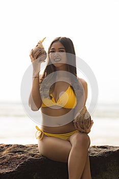 Young Asian woman sitting on the rock, holding coconuts. Summer beach concept. Tanned skin. Beautiful woman wearing yellow