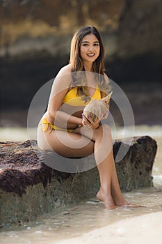 Young Asian woman sitting on the rock, holding coconuts. Summer beach concept. Tanned skin. Beautiful woman wearing yellow