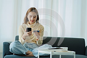 Young Asian woman sitting on sofa at home making online reservation on mobile phone