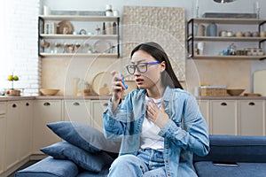 Young Asian woman sitting on sofa at home and using inhaler. Has an asthma attack, treats allergies