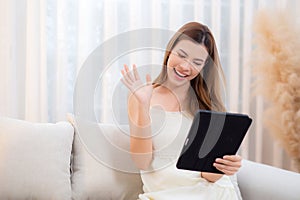 Young asian woman sitting on sofa using digital tablet computer for video call.