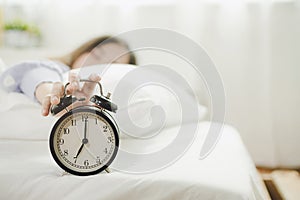 Young asian woman sleeping on bed pressing snooze button on black vintage alarm clock at seven o`clock morning in bed room at hom photo