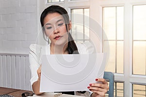 Young asian woman in smart casual wear holding document while sitting near window in retro home office