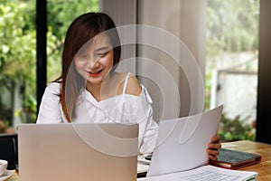 Young asian woman in smart casual wear holding document and working on laptop while sitting near window in home office