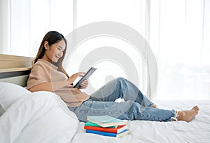 Young asian woman with a smile touching tablet computer screen in her hand while relax on bed at home, after reviewing the