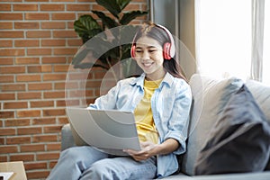 Young asian woman smiling listening to music using laptop while sitting on couch at home.
