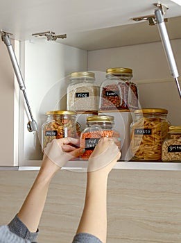Young Asian woman stacks glass jars with horns in kitchen cabinet. Storage of bulk products