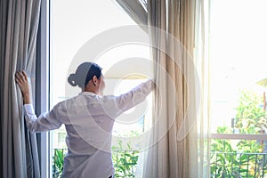 Young Asian woman standing and opening white curtain to looking outside in bedroom, female looking through the window in the