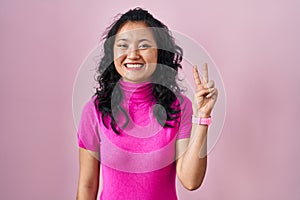 Young asian woman standing over pink background showing and pointing up with fingers number two while smiling confident and happy