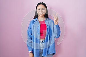 Young asian woman standing over pink background showing and pointing up with fingers number two while smiling confident and happy