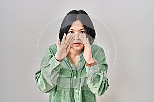 Young asian woman standing over white background hand on mouth telling secret rumor, whispering malicious talk conversation