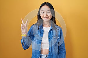 Young asian woman standing over yellow background showing and pointing up with fingers number two while smiling confident and
