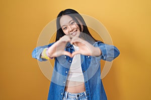 Young asian woman standing over yellow background smiling in love doing heart symbol shape with hands