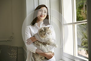 Young asian woman standying by window at home holding a cat
