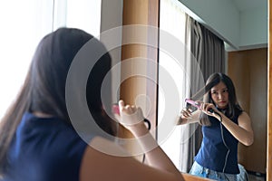 Young Asian woman straightening hair with hair straightener while looking into the mirror at home.