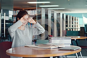 Young Asian woman student rubbing eyes, feeling tired after reading a book in library