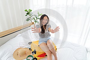 Young Asian woman in summer casual clothes is smiling and relaxing mood sitting on a yellow suitcase in her bedroom.