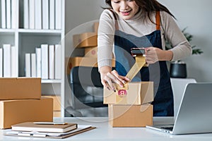 Young asian woman taping up a cardboard box in home office SME e-commerce business, relocation and new small business