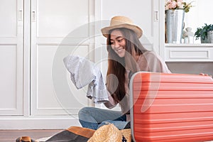 Young asian woman traveler prepare cloth into orange suitcase for holiday vacation at home.backpacker travel concept.