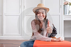 Young asian woman traveler thumbs up with orange suitcase prepare for holiday vacation at home.backpacker travel concept.