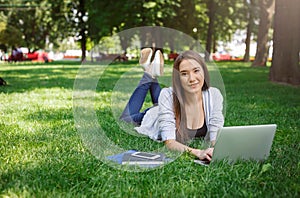 Young asian woman typing, laying on a green grass. Working with laptop in the park during the day