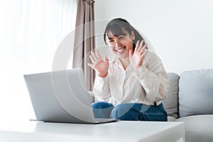 Young Asian woman using laptop computer for online video conference call waving hand making hello gesture on the couch in living