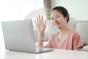 Young Asian woman using laptop computer for online video conference call waving hand making hello gesture in the living room