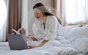 A young asian woman using laptop computer and working on paperwork while sitting on bed at home