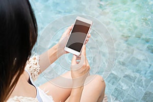 Young Asian Woman using mobile phone and sitting besides swimming pool. Summer vacation, technology and lifestyle concept