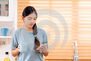 Young asian woman using smart phone with smiling, happy and relax emotion while drinking coffee in kitchen background, people on