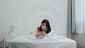 Young Asian woman using smartphone checking social media feeling happy smiling while lying on bed after wake up in the morning.