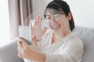 Young Asian woman using smartphone for online video conference call with friends waving hand making hello gesture
