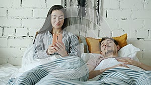 Young Asian woman using smartphone texting while husband sleeping in bed at home