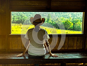 Young Asian woman wear the hat sit on wooden bench and watching beautiful view of green grass field and forest