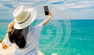 Young Asian woman wear straw hat in casual style use smartphone taking selfie at wooden pier. Summer vacation at tropical paradise