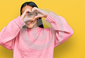 Young asian woman wearing casual winter sweater doing heart shape with hand and fingers smiling looking through sign