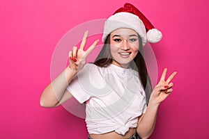 Young asian woman wearing christmas hat smiling with happy face winking at the camera doing victory sign isolated over pnk