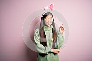 Young asian woman wearing cute easter bunny ears over pink background with a big smile on face, pointing with hand and finger to