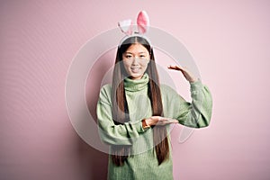 Young asian woman wearing cute easter bunny ears over pink background gesturing with hands showing big and large size sign,