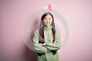 Young asian woman wearing cute easter bunny ears over pink background happy face smiling with crossed arms looking at the camera