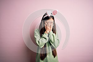 Young asian woman wearing cute easter bunny ears over pink background with sad expression covering face with hands while crying