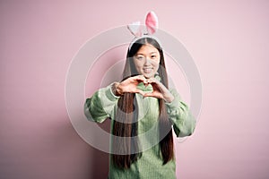 Young asian woman wearing cute easter bunny ears over pink background smiling in love doing heart symbol shape with hands