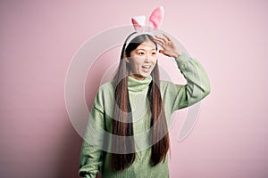 Young asian woman wearing cute easter bunny ears over pink background very happy and smiling looking far away with hand over head