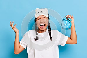 Young Asian woman wearing eye mask holding alarm clock screams from early wake up, with big smile and raised hands